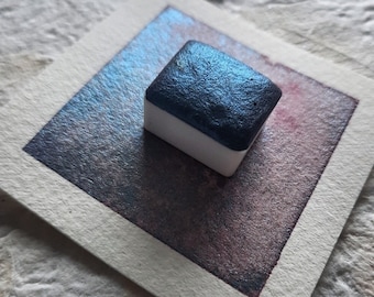 Pode incluir: Uma pastilha de aquarela quadrada com tinta metálica azul escura. A pastilha está sobre um quadrado de papel branco com uma lavagem de aquarela azul e roxa.