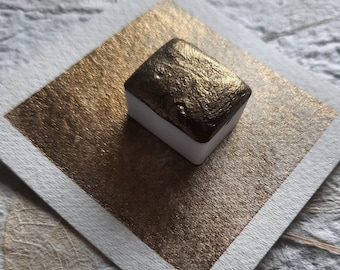 May include: A small, white pan of metallic gold watercolor paint. The paint is in a square pan and has a textured surface.