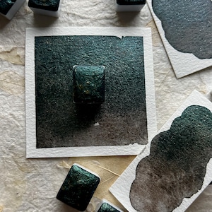 Puede incluir: Primer plano de pinturas de acuarela verde oscuro con motas doradas. Varios bloques de pintura rectangulares se encuentran sobre una superficie texturizada, junto con muestras en papel blanco. La pintura tiene un acabado brillante y met&aacute;lico.