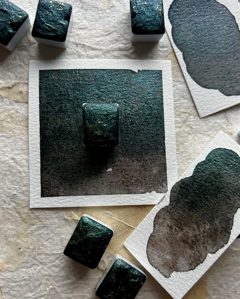 Puede incluir: Primer plano de pinturas de acuarela verde oscuro con motas doradas. Varios bloques de pintura rectangulares se encuentran sobre una superficie texturizada, junto con muestras en papel blanco. La pintura tiene un acabado brillante y met&aacute;lico.