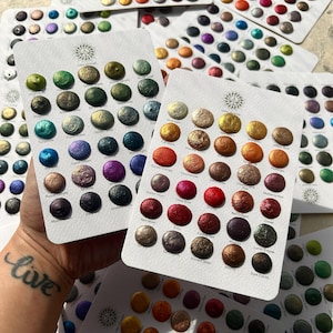 May include: A collection of watercolor paint swatches in various colors, arranged on white cards. Each card features multiple circular paint samples, showcasing a range of metallic and iridescent hues. The cards are held in a hand, with other cards in the background.