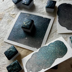 Puede incluir: Una colecci&oacute;n de pinturas de acuarela en verde azulado oscuro y dorado, con varios bloques de pintura cuadrados y muestras en papel blanco. La pintura tiene un acabado brillante y met&aacute;lico, y las muestras muestran el degradado del color.