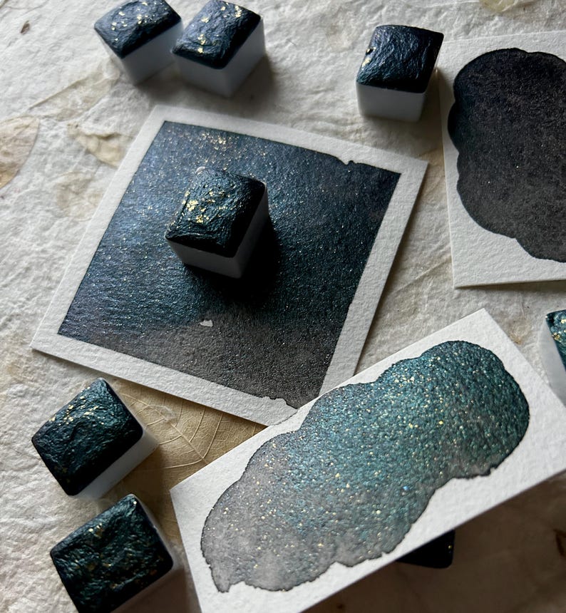 Puede incluir: Una colecci&oacute;n de pinturas de acuarela en verde azulado oscuro y dorado, con varios bloques de pintura cuadrados y muestras en papel blanco. La pintura tiene un acabado brillante y met&aacute;lico, y las muestras muestran el degradado del color.
