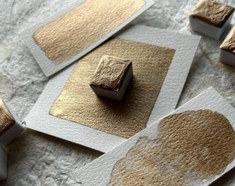 May include: Four small pans of metallic gold watercolor paint on white paper squares. The pans are arranged in a square shape with one pan in the center.