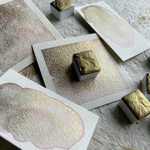Può includere: Primo piano di pastiglie di vernice ad acquerello con finitura oro metallizzato. Le pastiglie sono disposte su carta bianca con campioni di vernice dorata.