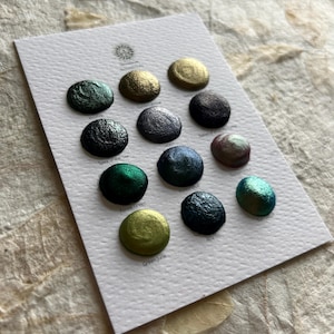 May include: A white card showcases twelve metallic watercolour paint samples in shades of green, gold, blue, and purple. The paints are arranged in a grid, each labelled with a unique name. The card rests on a textured, light-coloured surface.