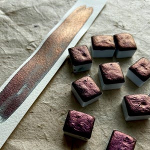 May include: Close-up of watercolor paints in small square pans. The paints are a deep, shimmering burgundy color. A swatch of the same color is on a white strip of paper. The background is a textured, light brown surface.
