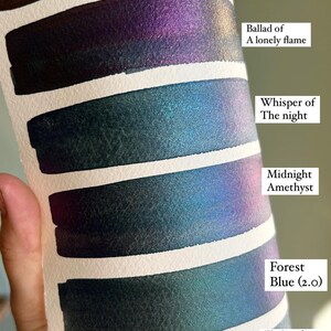 May include: A watercolor swatch card featuring six color samples with metallic shimmer. The colors are labeled "Garnet Sunset", "Ballad of A lonely flame", "Whisper of The night", "Midnight Amethyst", "Forest Blue (2.0)", and "Flight of The Dragonflies".