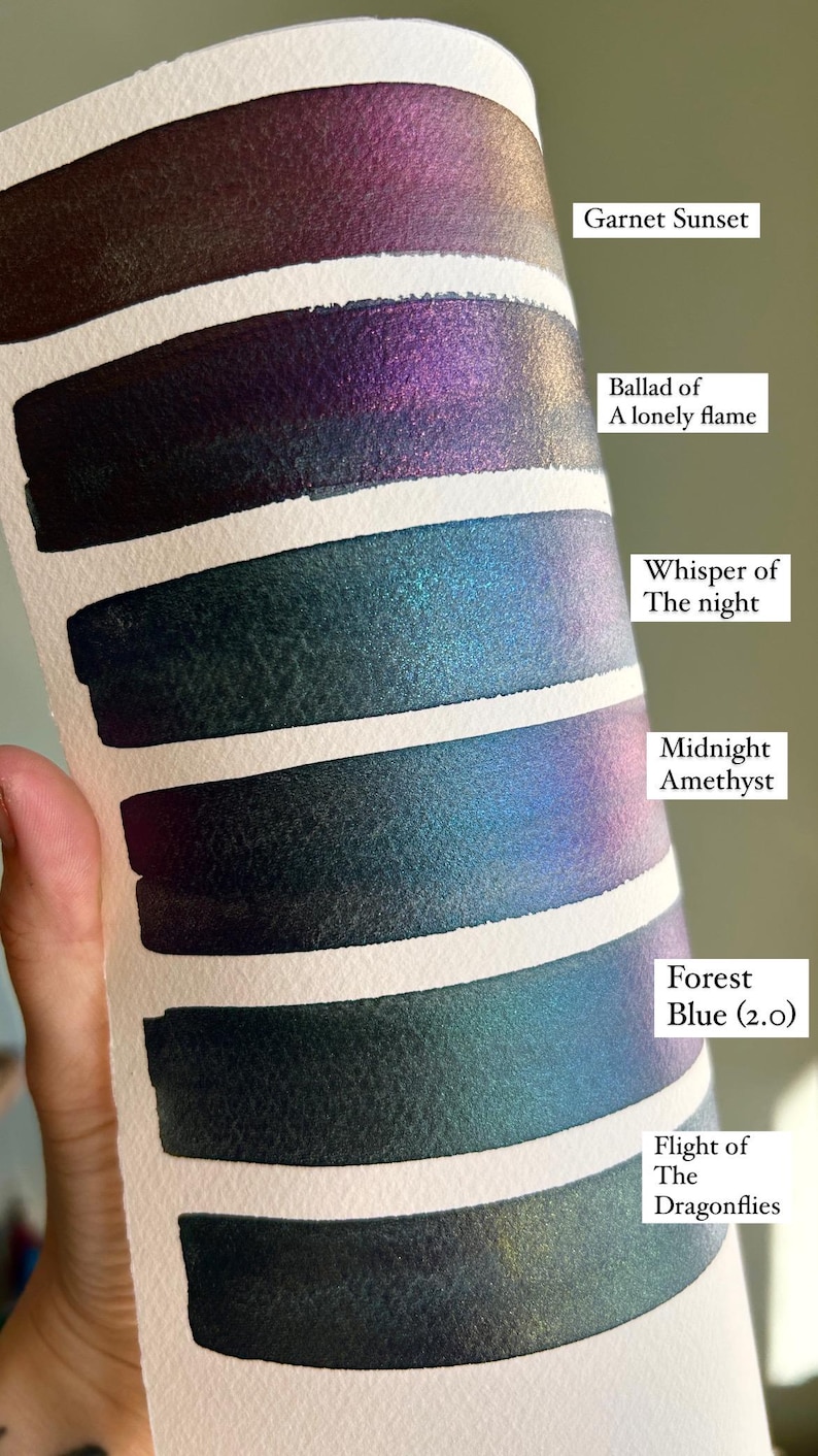 May include: A watercolor swatch card featuring six color samples with metallic shimmer. The colors are labeled "Garnet Sunset", "Ballad of A lonely flame", "Whisper of The night", "Midnight Amethyst", "Forest Blue (2.0)", and "Flight of The Dragonflies".