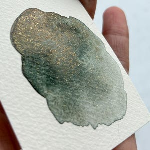 May include: A close-up of a dark green watercolor wash with gold glitter. The wash is on a white textured paper.