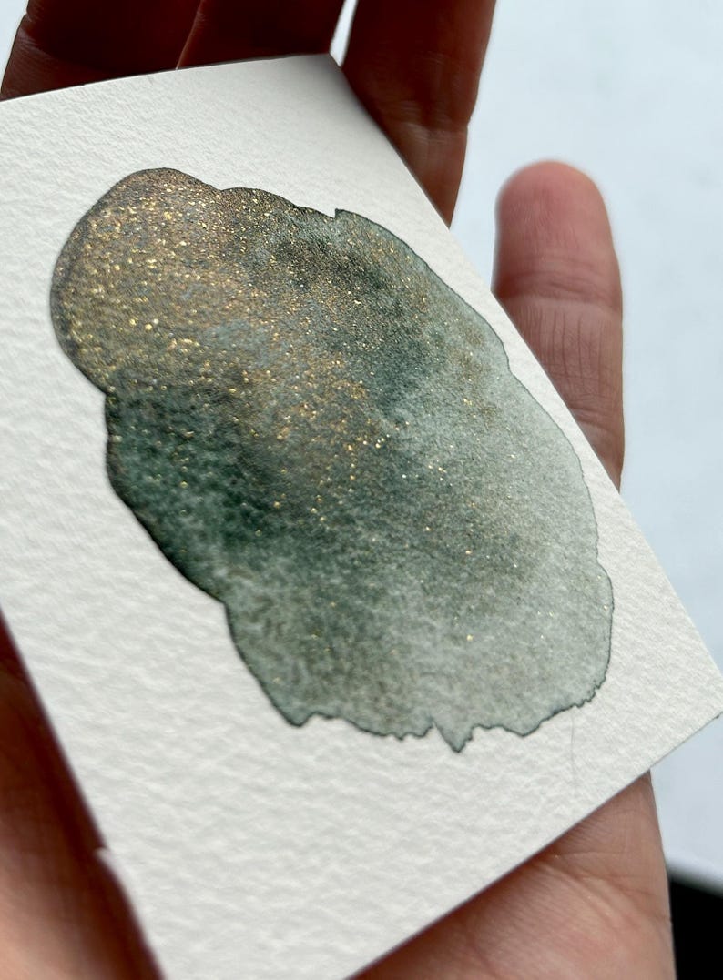 May include: A close-up of a dark green watercolor wash with gold glitter. The wash is on a white textured paper.