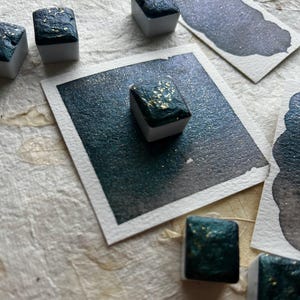Puede incluir: Primer plano de cubos de pintura de acuarela con un tono verde azulado oscuro y motas doradas. Varias muestras del mismo color se muestran sobre papel de acuarela blanco. Los cubos de pintura est&aacute;n sobre una superficie texturizada blanquecina.
