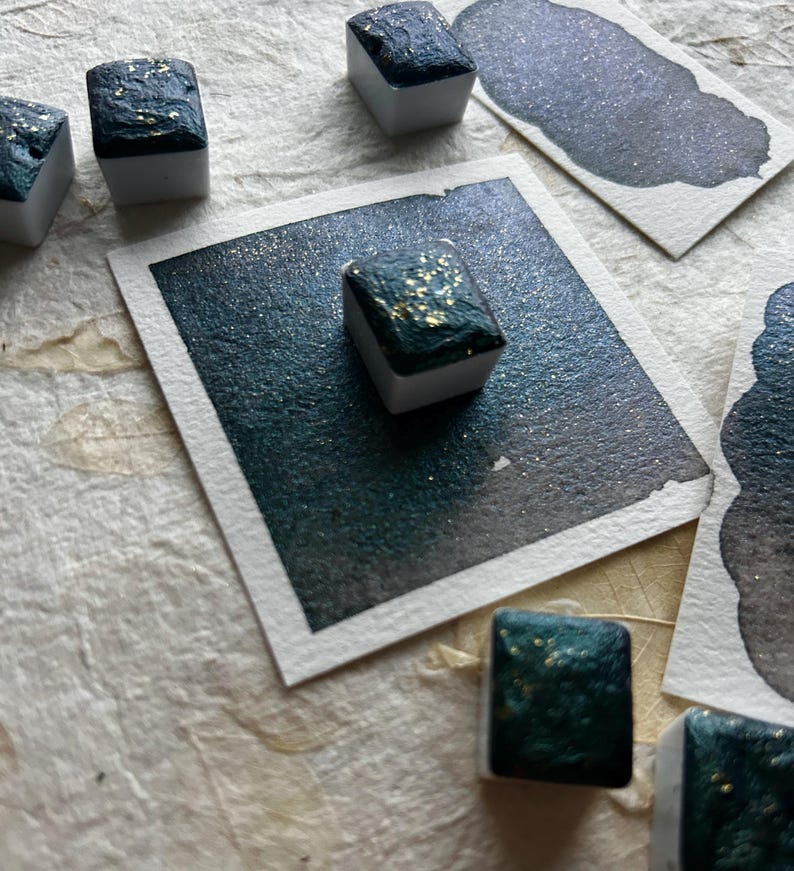 Puede incluir: Primer plano de cubos de pintura de acuarela con un tono verde azulado oscuro y motas doradas. Varias muestras del mismo color se muestran sobre papel de acuarela blanco. Los cubos de pintura est&aacute;n sobre una superficie texturizada blanquecina.