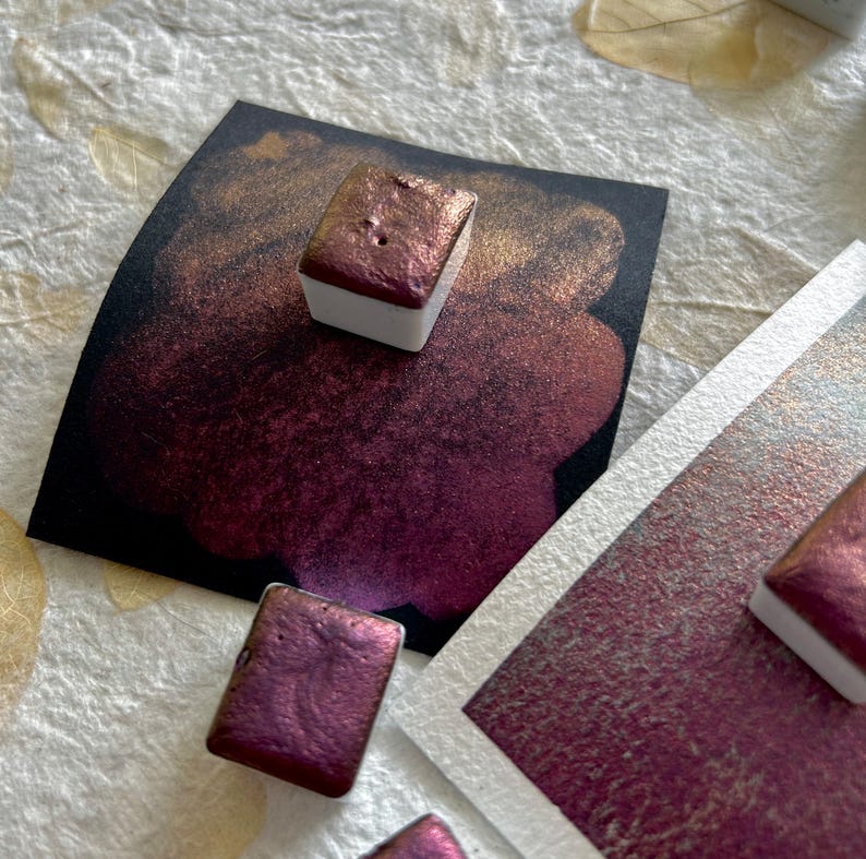 May include: Close-up of watercolor paints in a variety of shades of purple and gold. The paints are in small, square pans with white bases. The colors are swatched on black and white paper, showing their metallic sheen.
