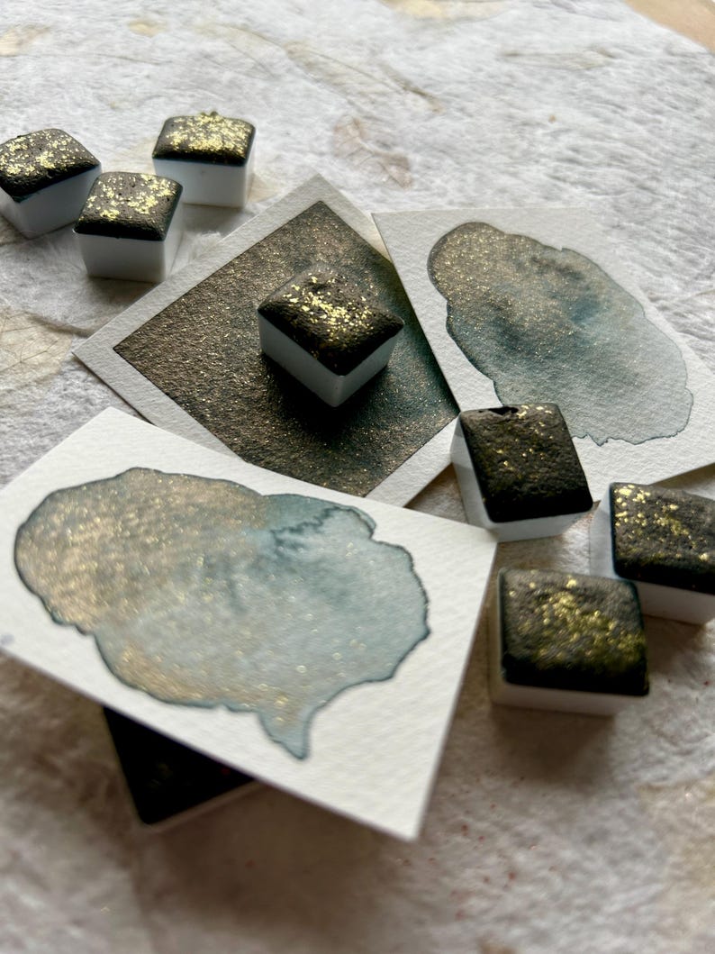 May include: A set of eight watercolor paint pans with a gold shimmer. The pans are arranged in a semi-circle on a white surface. The paint is a dark green with gold flecks.