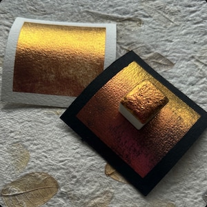 Può includere: Due campioni di pittura ad acquerello con finitura oro metallizzato. Uno è un quadrato di vernice dorata su carta bianca, l'altro è un quadrato di vernice dorata su carta nera. Un piccolo quadrato di vernice dorata è su una base di ceramica bianca.