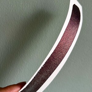 May include: A curved swatch of watercolor paint on white paper. The paint transitions from a deep burgundy to a lighter, shimmering red-violet hue. The paper is held against a muted green background.