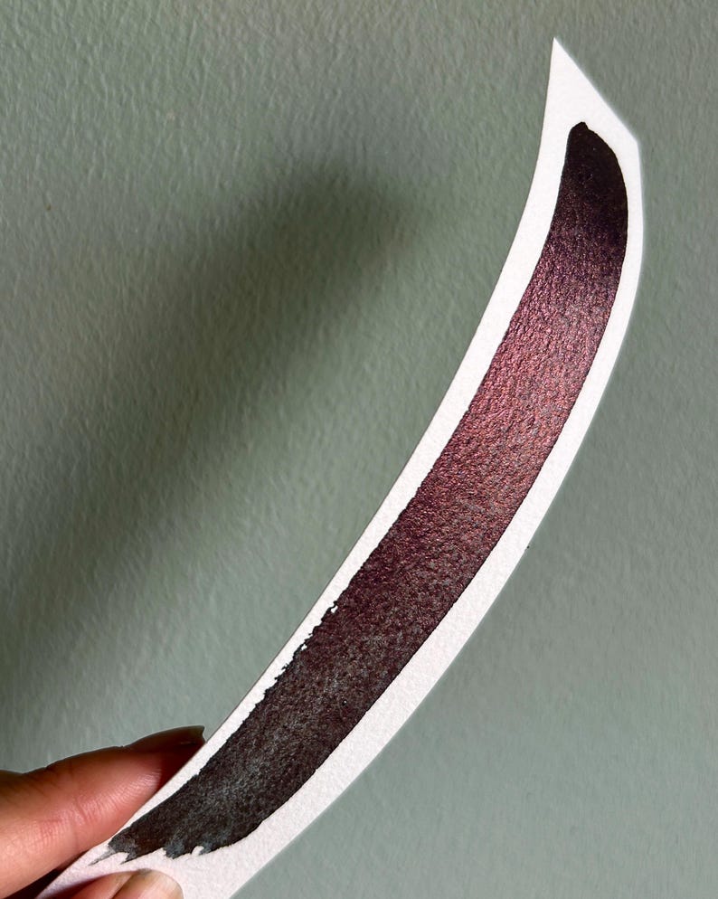 May include: A curved swatch of watercolor paint on white paper. The paint transitions from a deep burgundy to a lighter, shimmering red-violet hue. The paper is held against a muted green background.