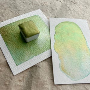 Può includere: Un piccolo panetto di acquerello verde con un effetto metallico. Il colore è mostrato su un cartoncino bianco con un campione del colore dipinto accanto.