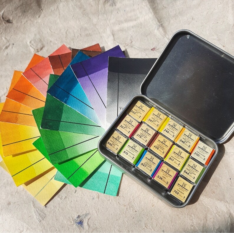 Puede incluir: Un juego de 16 acuarelas en una lata de metal. Las pinturas est&aacute;n dispuestas en una cuadr&iacute;cula y est&aacute;n etiquetadas con n&uacute;meros. La lata est&aacute; abierta y las pinturas son visibles. Los colores de las pinturas son una mezcla de tonos c&aacute;lidos y fr&iacute;os.