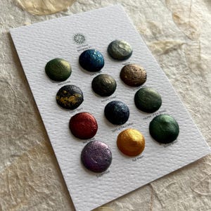 Peut inclure: Une carte blanche présente douze échantillons de peinture aquarelle aux couleurs variées, dont du vert, du bleu, du rouge et de l'or. Chaque échantillon est un petit point rond avec un éclat métallique, nommé "Kelp Forest" et "Full Moon".