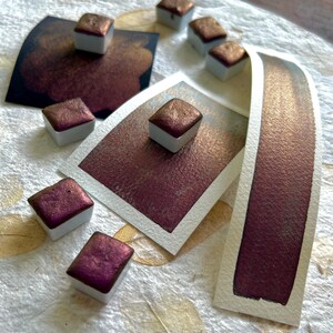 May include: Close-up of watercolor paints in square pans with a metallic, reddish-brown hue. Several swatches of the same color are displayed on white paper, showcasing the paint's shimmer and depth. The art supplies are arranged on a textured, off-white surface.