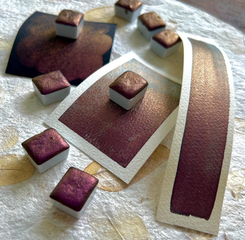 May include: Close-up of watercolor paints in square pans with a metallic, reddish-brown hue. Several swatches of the same color are displayed on white paper, showcasing the paint's shimmer and depth. The art supplies are arranged on a textured, off-white surface.