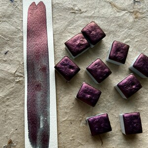 May include: A swatch of metallic burgundy watercolor paint next to several square pans of the same color. The paint is swatched on a white strip of paper, showing a gradient from dark to light. The pans are on a textured, tan surface.