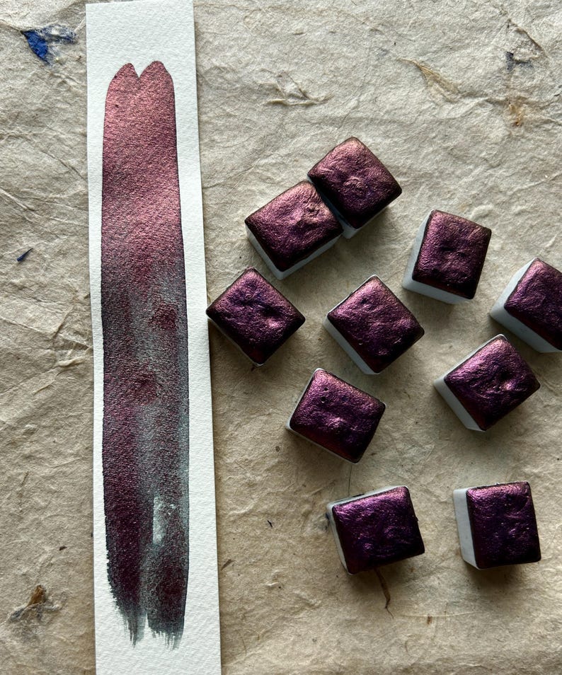May include: A swatch of metallic burgundy watercolor paint next to several square pans of the same color. The paint is swatched on a white strip of paper, showing a gradient from dark to light. The pans are on a textured, tan surface.