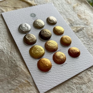 May include: A white card displays a collection of metallic watercolor paints in various shades of gold, bronze, and silver. The colors include "Silver Gold", "Payne", "Green Gold", "Manganese", "Silk Gold", "New Moon", "Golden Chrysanthemum", "Honey", "Full Moon", and "Imperial Gold".