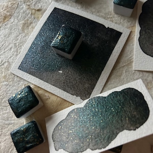 Puede incluir: Primer plano de pinturas de acuarela en tonos verde azulado y dorado. Las pinturas est&aacute;n en peque&ntilde;os recipientes cuadrados, con un degradado de color en el papel. La imagen muestra una variedad de pinceladas y muestras de color, resaltando el brillo de la pintura.