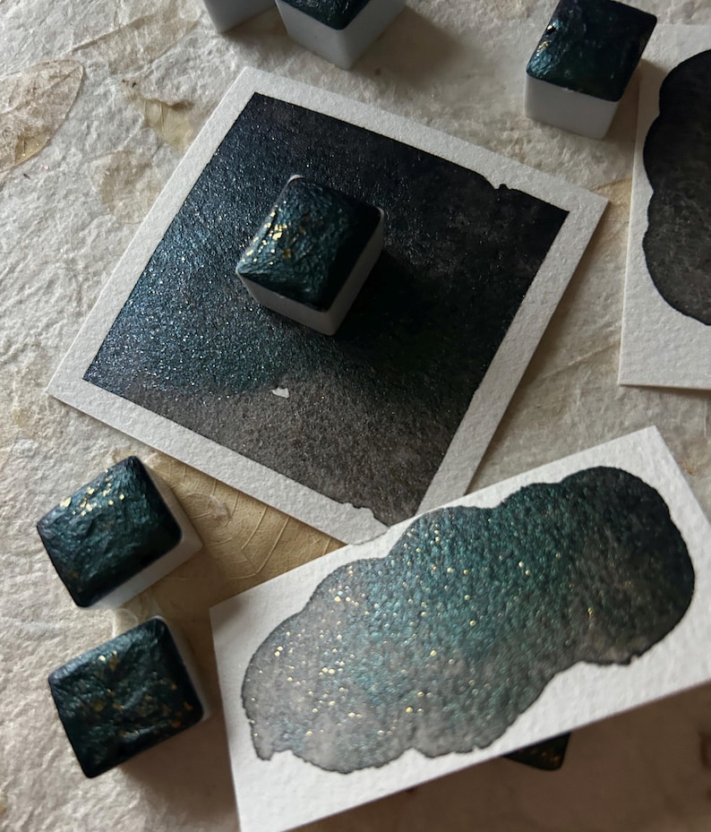 Puede incluir: Primer plano de pinturas de acuarela en tonos verde azulado y dorado. Las pinturas est&aacute;n en peque&ntilde;os recipientes cuadrados, con un degradado de color en el papel. La imagen muestra una variedad de pinceladas y muestras de color, resaltando el brillo de la pintura.