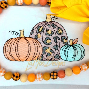 May include: Three embroidered pumpkins in different styles. The largest pumpkin is purple with a leopard print, the middle pumpkin is orange, and the smallest pumpkin is light blue.