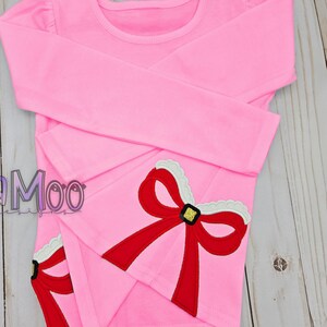 May include: Pink long-sleeved shirt with a red bow and gold trim on the sleeve. The bow has white trim around the edges.