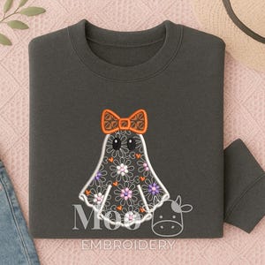 May include: Dark grey sweatshirt with a ghost design, orange bow, and floral pattern. The ghost has large eyes and is decorated with white, purple, and orange flowers. The sweatshirt is folded on a pink surface.