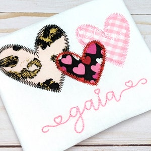 May include: Three embroidered hearts in different patterns, including leopard print, pink hearts on black, and pink and white gingham. The word "gala" is embroidered in pink cursive below the hearts.