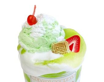 May include: A dessert parfait in a clear glass, featuring layers of green and white cream, topped with a scoop of green ice cream, a red cherry, a strawberry slice, and a cookie with the word "CHOICE". The glass has the words "MELON PARFAIT".