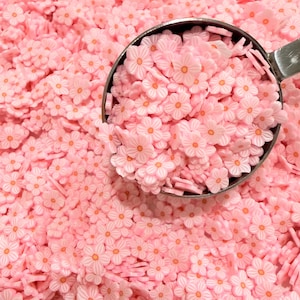 Pode incluir: Uma imagem de perto de uma pilha de pequenas decorações em forma de flor rosa. Uma concha de metal contém uma porção das flores, mostrando seu design delicado com pétalas brancas e centros laranja. O fundo é preenchido com os mesmos enfeites florais.