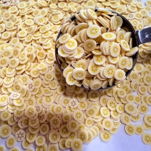 Pode incluir: Close-up de inúmeras pequenas fatias de banana, cada uma com um centro amarelo e um anel externo branco. Uma concha de metal contém uma porção das fatias. As fatias estão espalhadas sobre uma superfície branca. As fatias de banana são um elemento decorativo.