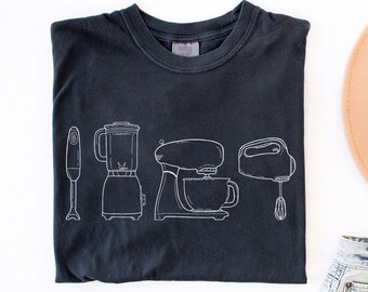 May include: A black t-shirt featuring white line drawings of kitchen appliances: an immersion blender, a blender, a stand mixer, and a hand mixer. The image also includes a tan hat, denim shorts, and gold-toned sandals.