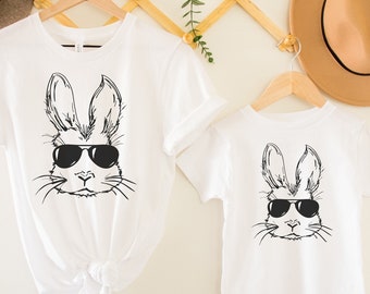 Peut inclure: Deux t-shirts blancs avec une illustration en noir et blanc d'un lapin portant des lunettes de soleil. Les t-shirts sont suspendus à des cintres contre un mur blanc.