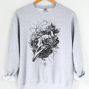 May include: Grey sweatshirt with a black and white graphic print of a skull with a geometric design and floral elements.