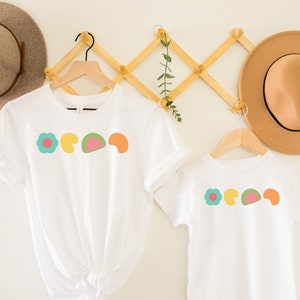 May include: Two white t-shirts with a colorful flower and geometric shapes design. The shirts are hanging on a wooden rack with hats.