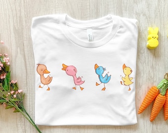 Peut inclure: T-shirt blanc avec un motif de cinq canards de dessin animé en ligne. Les canards sont de couleurs différentes : marron, rose, bleu, jaune et marron. Les canards sont tous tournés vers la droite et ont les ailes déployées.