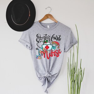 May include: A gray t-shirt with the text "Santa's Favorite Nurse" in black and red lettering. The design includes a nurse's cap, stethoscope, bandages, a cup of coffee, and a Christmas stocking.