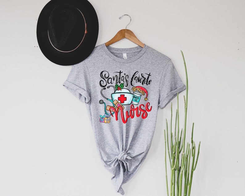 May include: A gray t-shirt with the text "Santa's Favorite Nurse" in black and red lettering. The design includes a nurse's cap, stethoscope, bandages, a cup of coffee, and a Christmas stocking.