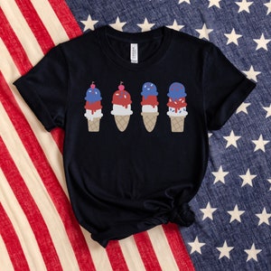 May include: A black t-shirt featuring a design of four ice cream cones. Each cone has three scoops of ice cream in red, white, and blue, topped with a cherry. The ice cream is dripping down the sides of the cones.