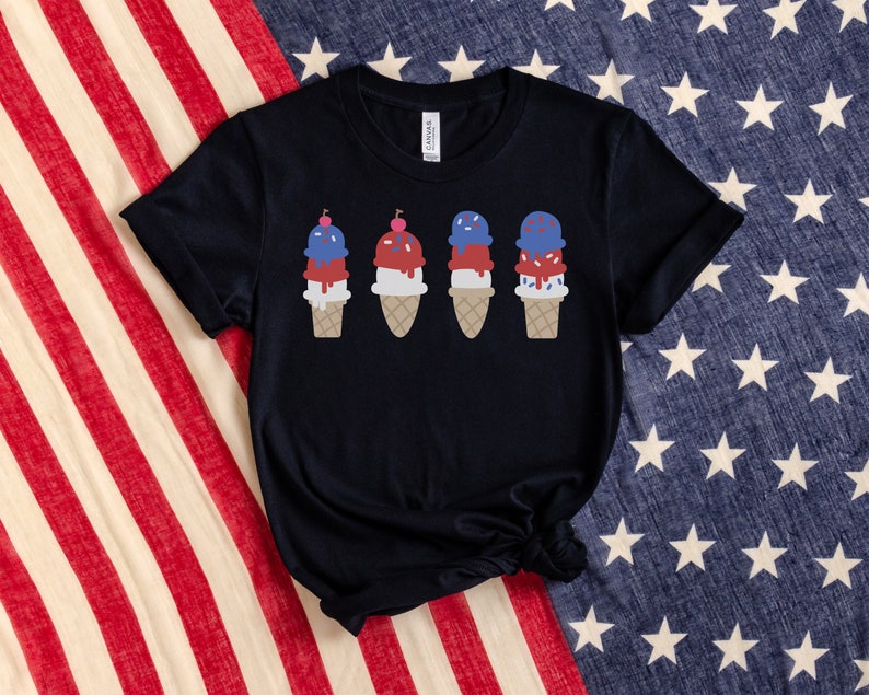 May include: A black t-shirt featuring a design of four ice cream cones. Each cone has three scoops of ice cream in red, white, and blue, topped with a cherry. The ice cream is dripping down the sides of the cones.
