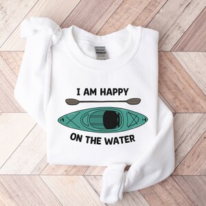 May include: White sweatshirt with the text "I am happy on the water" and a green kayak with a brown paddle.