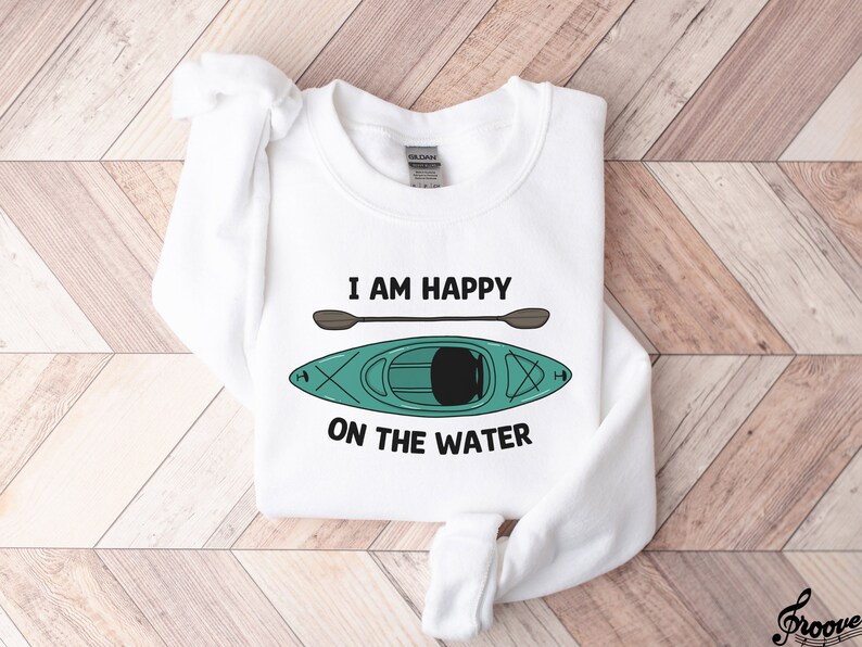 May include: White sweatshirt with the text "I am happy on the water" and a green kayak with a brown paddle.